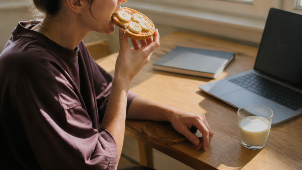 Woman eating peanut butter and banana toast with milk at a home office desk.