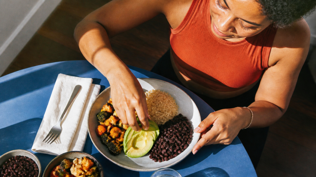 Black woman arranging avocado on a plate with roasted vegetables, black beans, and brown rice.