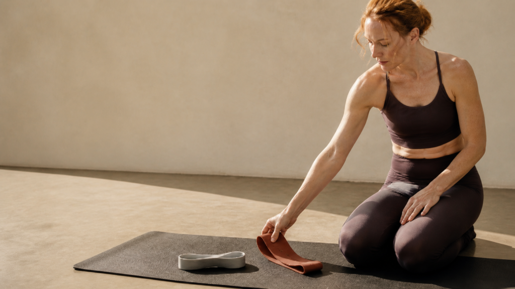Red-haired woman reaching for a terracotta resistance band beside a light dumbbell on an exercise mat.